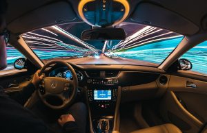 Tesla Robotaxi Passengers Report Lane Drifting, Braking A Tesla robotaxi with a human safety monitor in the front seat drives on an Austin street, surrounded by traffic and pedestrians.